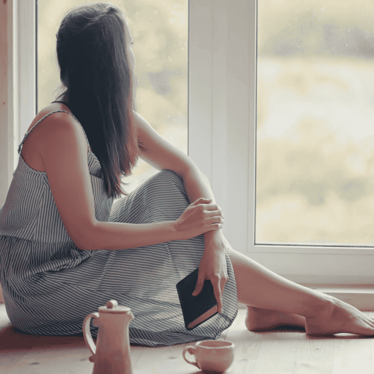 A reflective woman gazing out the window with a book in hand, symbolizing emotional depth and wondering if the her symptoms she is experience are signs of an empath.